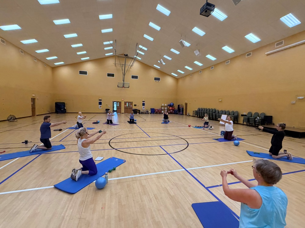 The interior of a gymnasium with a class of people sitting on yoga mats on the floor, with weights next to them, and stretching
